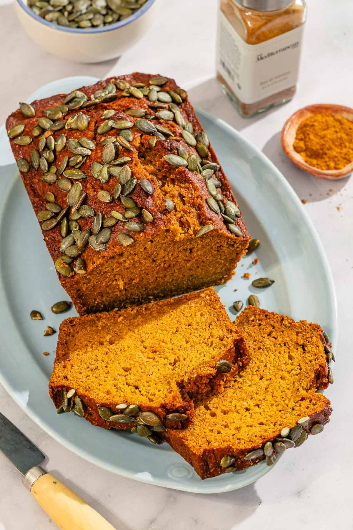 A partial pumpkin bread loaf along with 2 slices on a platter. Next to this is a knife, a bottle of ras el hanout, and bowls of pumpkin seeds and ras el hanout.
