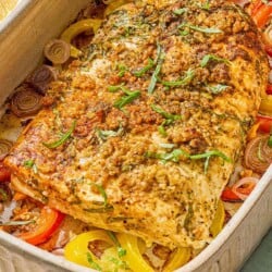 The baked fish recipe on a bed of peppers and shallots in a baking dish next to two lemon halves.