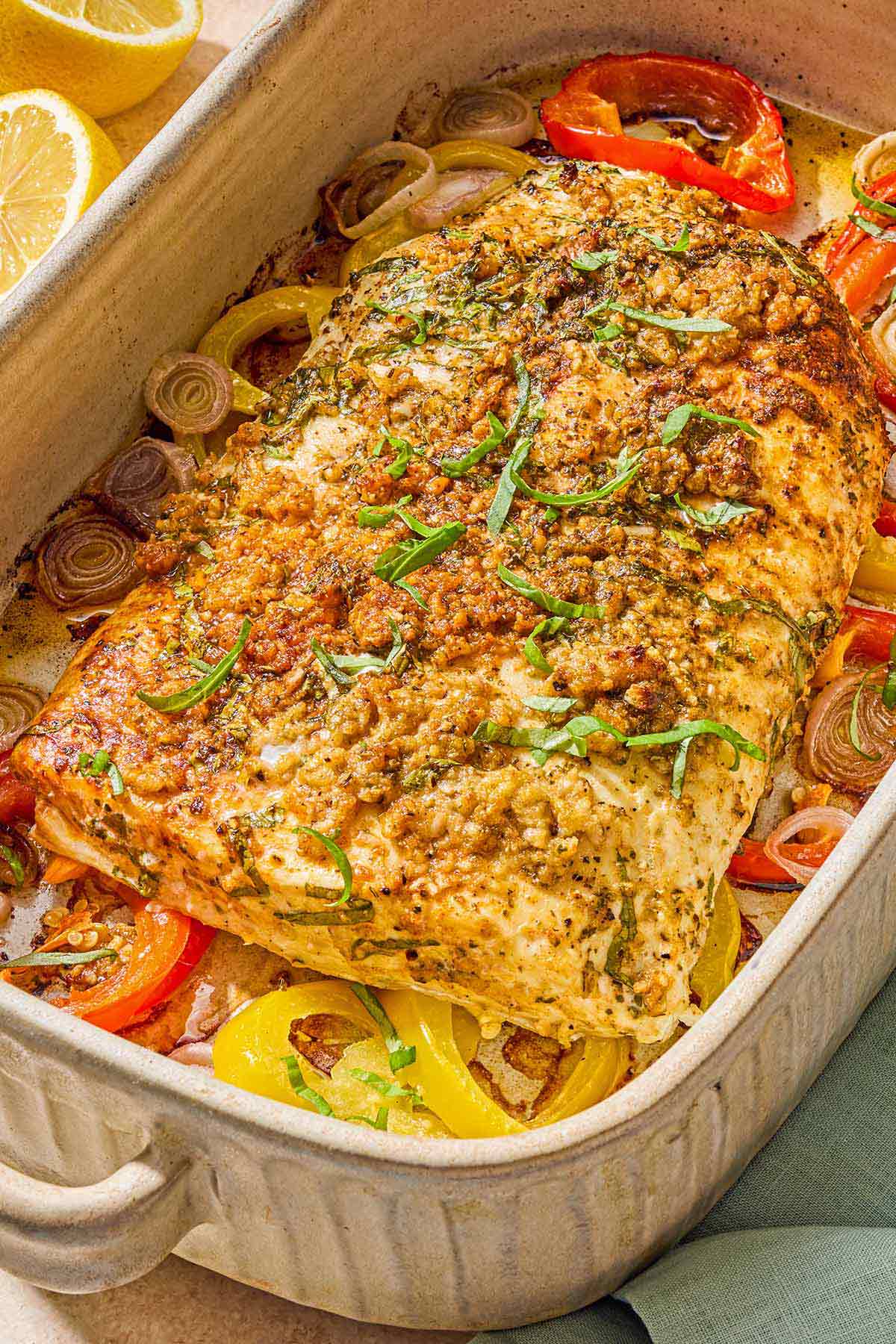 The baked fish recipe on a bed of peppers and shallots in a baking dish next to two lemon halves.