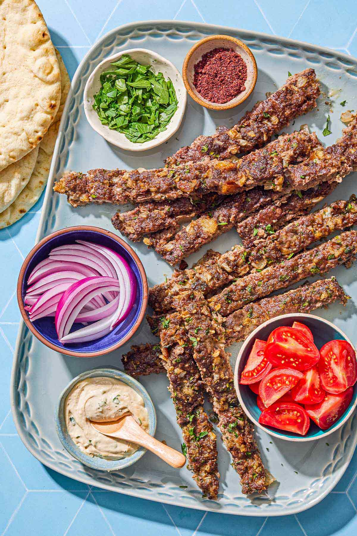 The pieces of the sheet pan baked kofta on a platter with bowls of red onion, tomatoes, sumac, parsley and tahini. Next to this is a stack of pita bread.