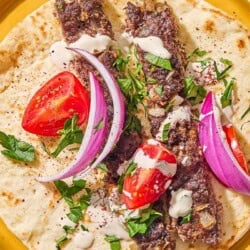 Two pieces of the sheet pan baked kofta on a piece of pita topped with parsley, red onion, tomato and tahini on a plate.