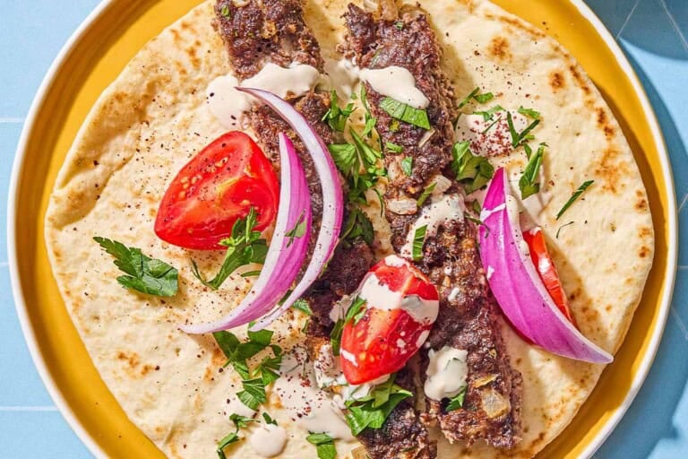 Two pieces of the sheet pan baked kofta on a piece of pita topped with parsley, red onion, tomato and tahini on a plate.