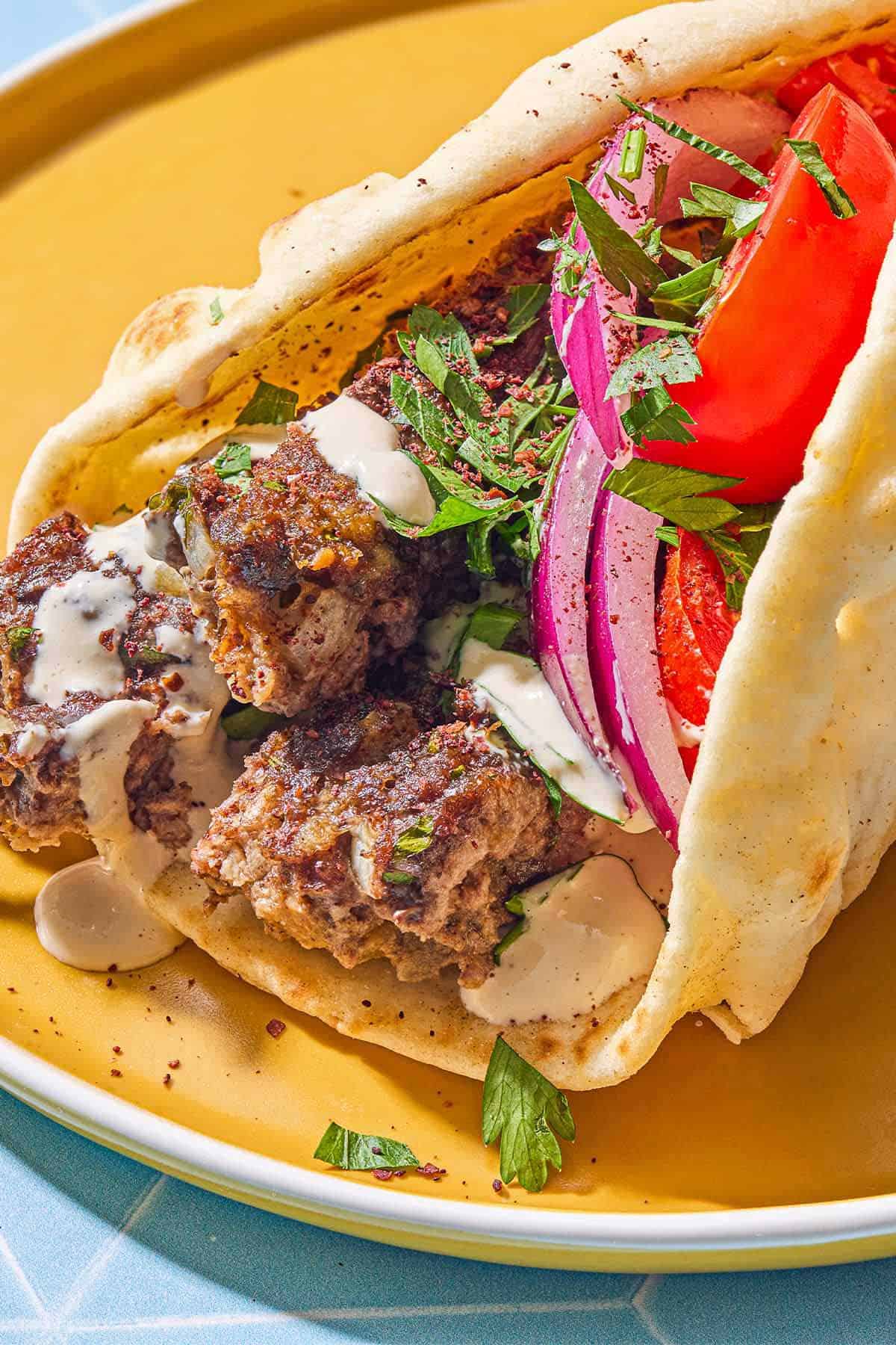 A pita wrapped filled with the sheet pan baked kofta topped with red onion, tomatoes, parsley and tahini on a plate.