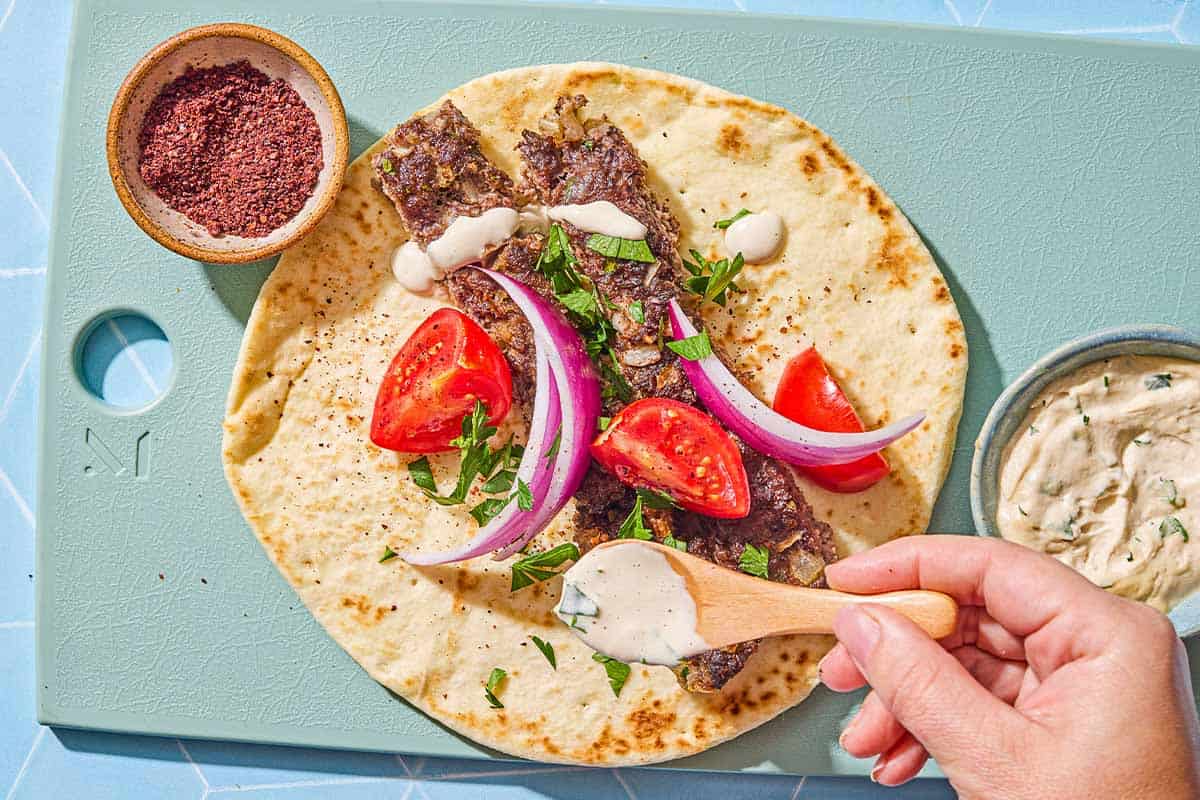 Two pieces of the sheet pan baked kofta on a piece of pita topped with parsley, red onion and tomato being drizzled with tahini on a cutting board. Next to this is a bowl of tahini and a bowl of sumac.