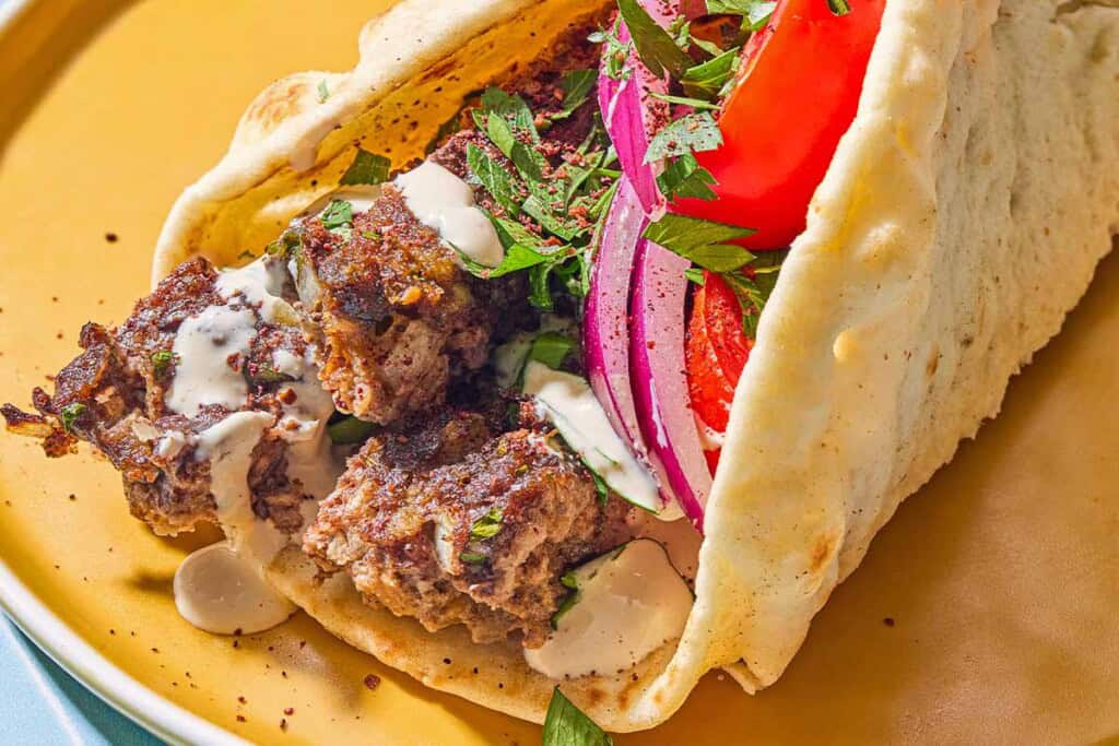A pita wrapped filled with the sheet pan baked kofta topped with red onion, tomatoes, parsley and tahini on a plate.