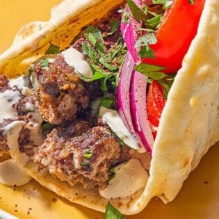A pita wrapped filled with the sheet pan baked kofta topped with red onion, tomatoes, parsley and tahini on a plate.