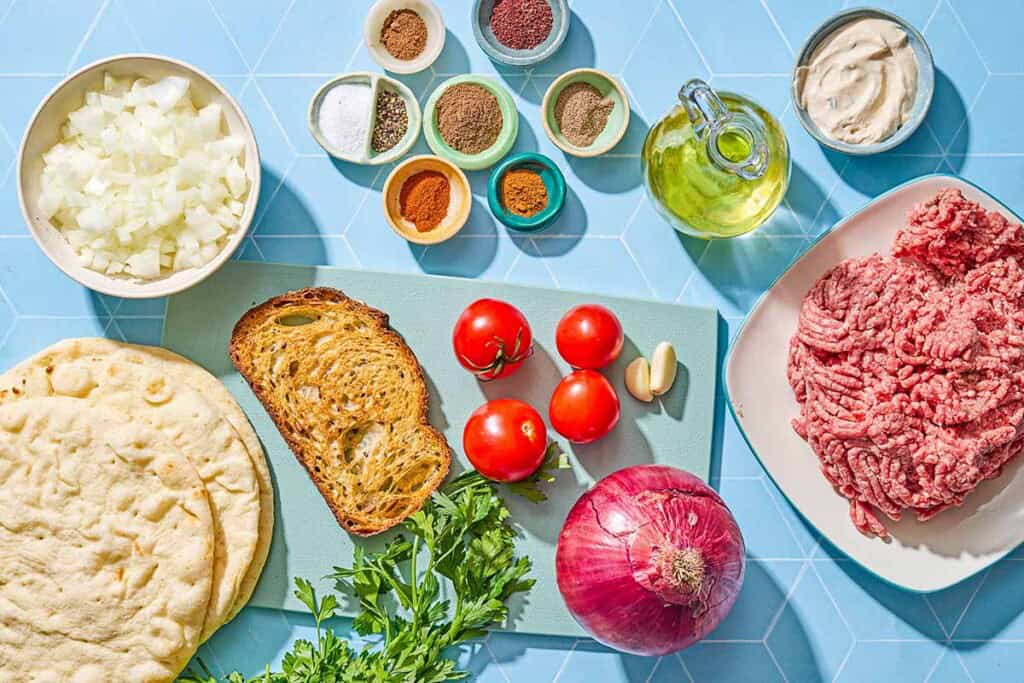 Ingredients for sheet pan baked kofta including ground beef, a slice of bread, onion, garlic, parsley, salt, pepper, allspice, cayenne, cardamom, sumac, nutmeg, paprika, olive oil, pita bread, tahini sauce, tomatoes, and red onion.