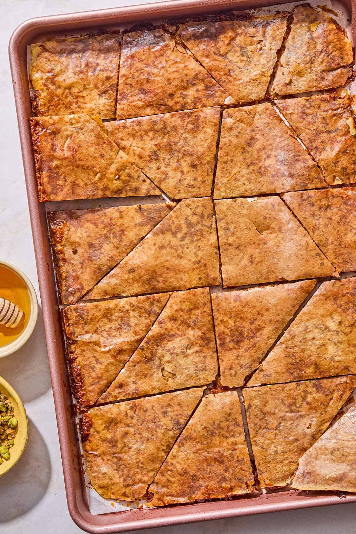 Broken pieces of baklava bark on a parchment lined sheet pan. Next to this are small bowls of chopped pistachios and honey.