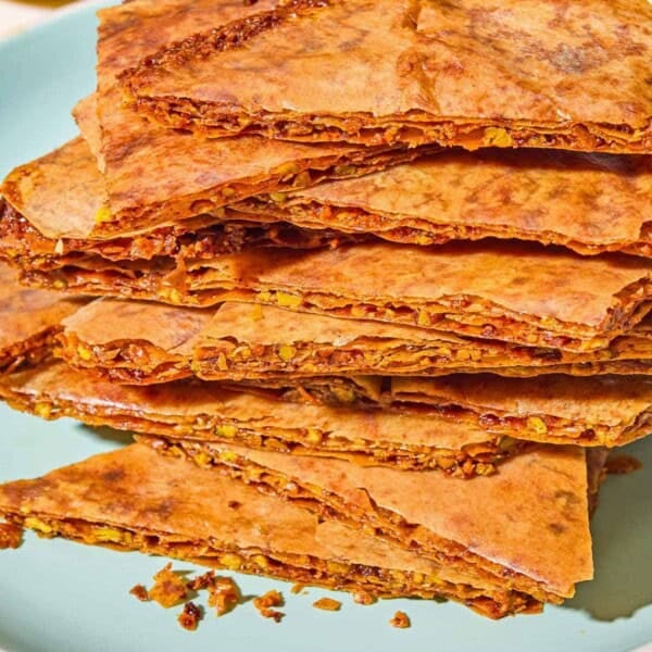 A close up of a stack of baklava bark on a plate. In the background are bowls of chopped pistachios and honey.