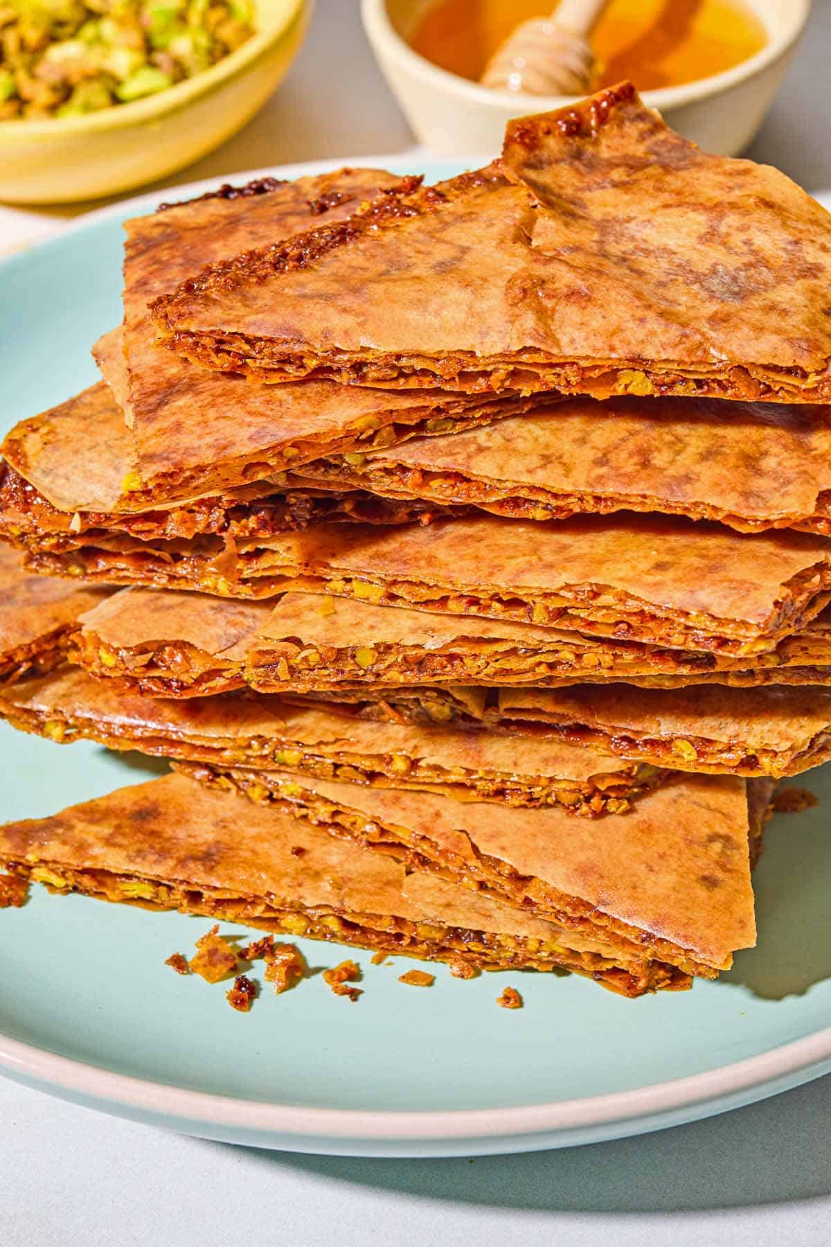A close up of a stack of baklava bark on a plate. In the background are bowls of chopped pistachios and honey.