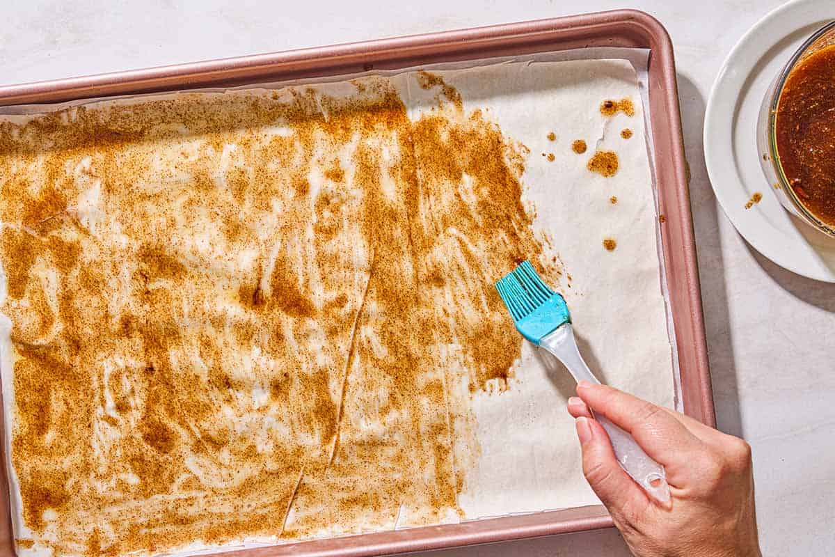 The honey mixture for the baklava bark being brushed on a sheet of phyllo dough lining a sheet pan. Next to this is the rest of the honey mixture in a bowl.