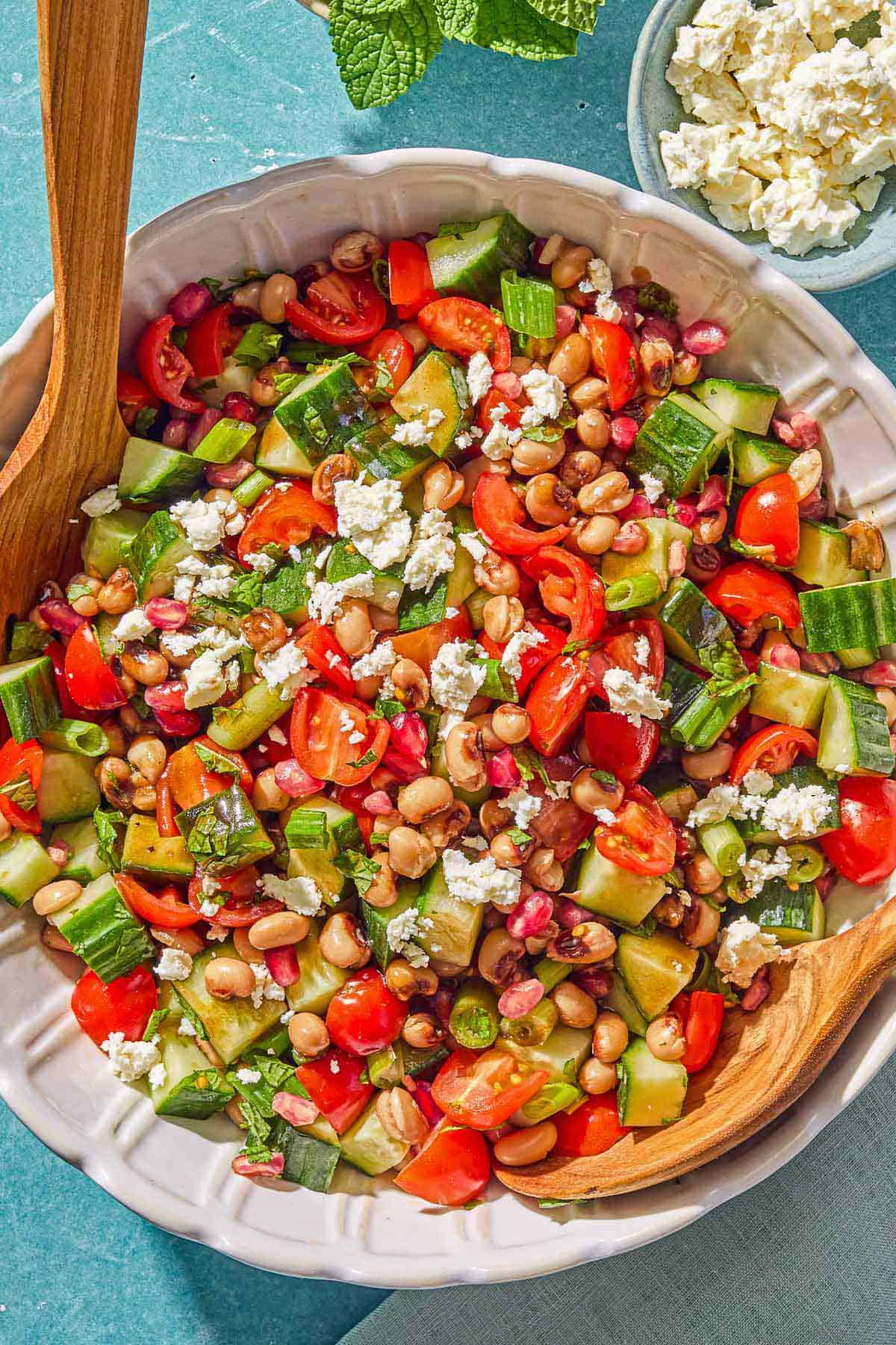 The black eyed pea salad in a bowl with wooden serving utensils. Next to this are bowls of mint and crumbled feta.