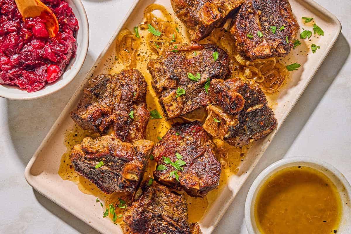 Several braised lamb chops on a platter. Next to this are bowls of the braising sauce and the cranberry harissa chutney.