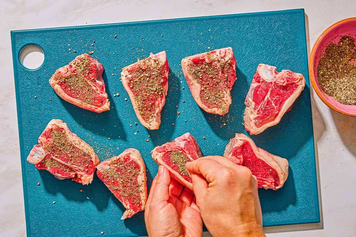Several seasoned raw lamb shanks on a cutting board, with one being coated in the seasoning. Next to this is a bowl of the seasoning.