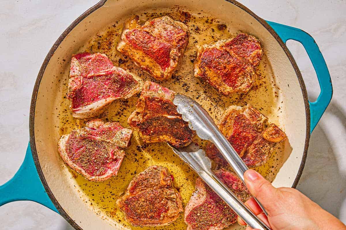 The seasoned lamb chopped being browned in the braiser. One is being flipped with a pair of tongs.