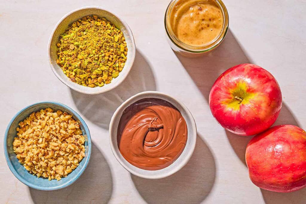 Ingredients for the caramel apple slices including apples, date caramel sauce, melted chocolate, chopped pistachios and chopped walnuts.