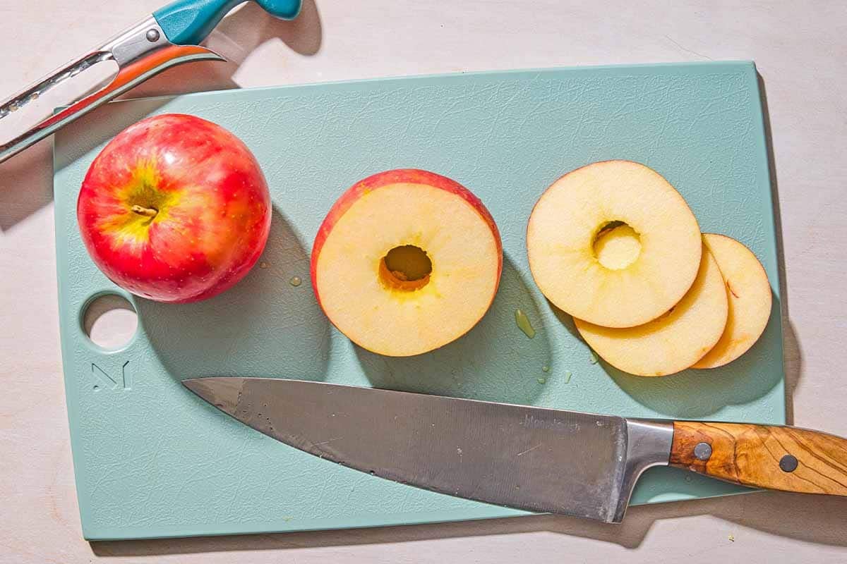 Several apple rings on a cutting board with a whole apple and a knife. Next to this is an apple corer.
