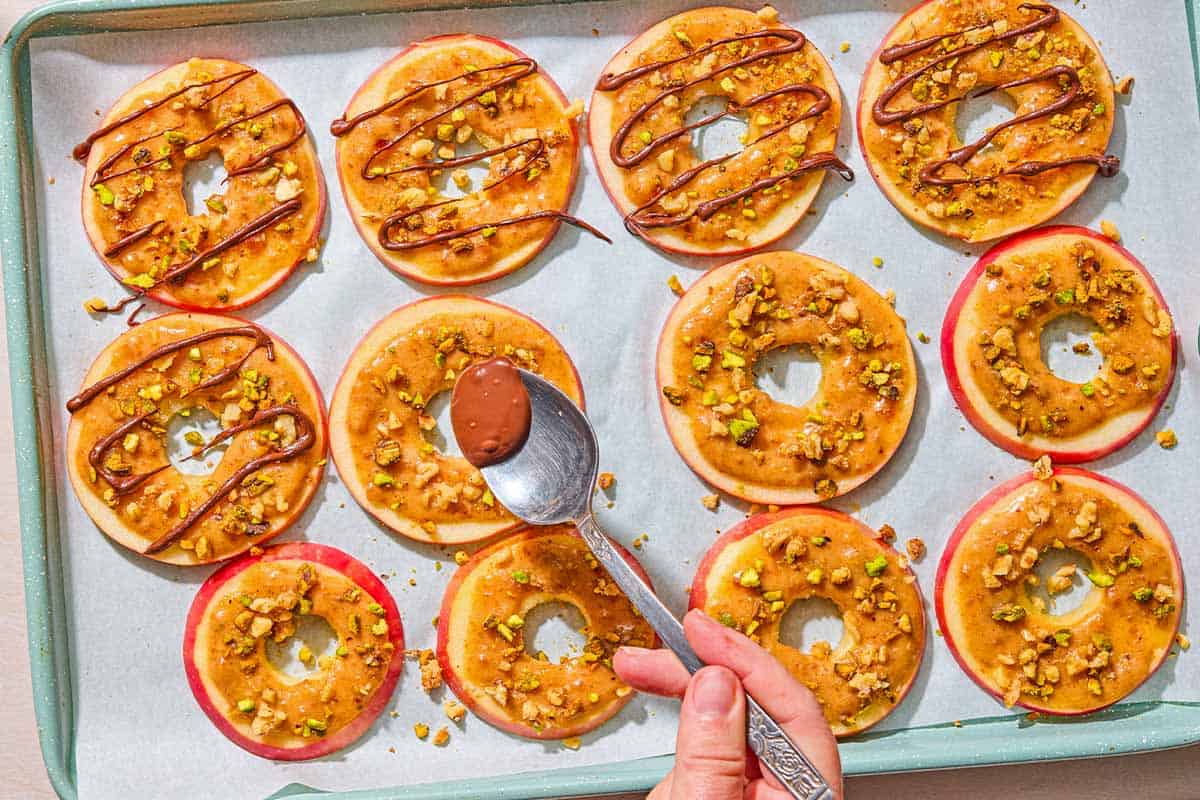 Chocolate being drizzled onto a caramel apple slice that is on a parchment lined sheet pan with several other caramel apple slices.