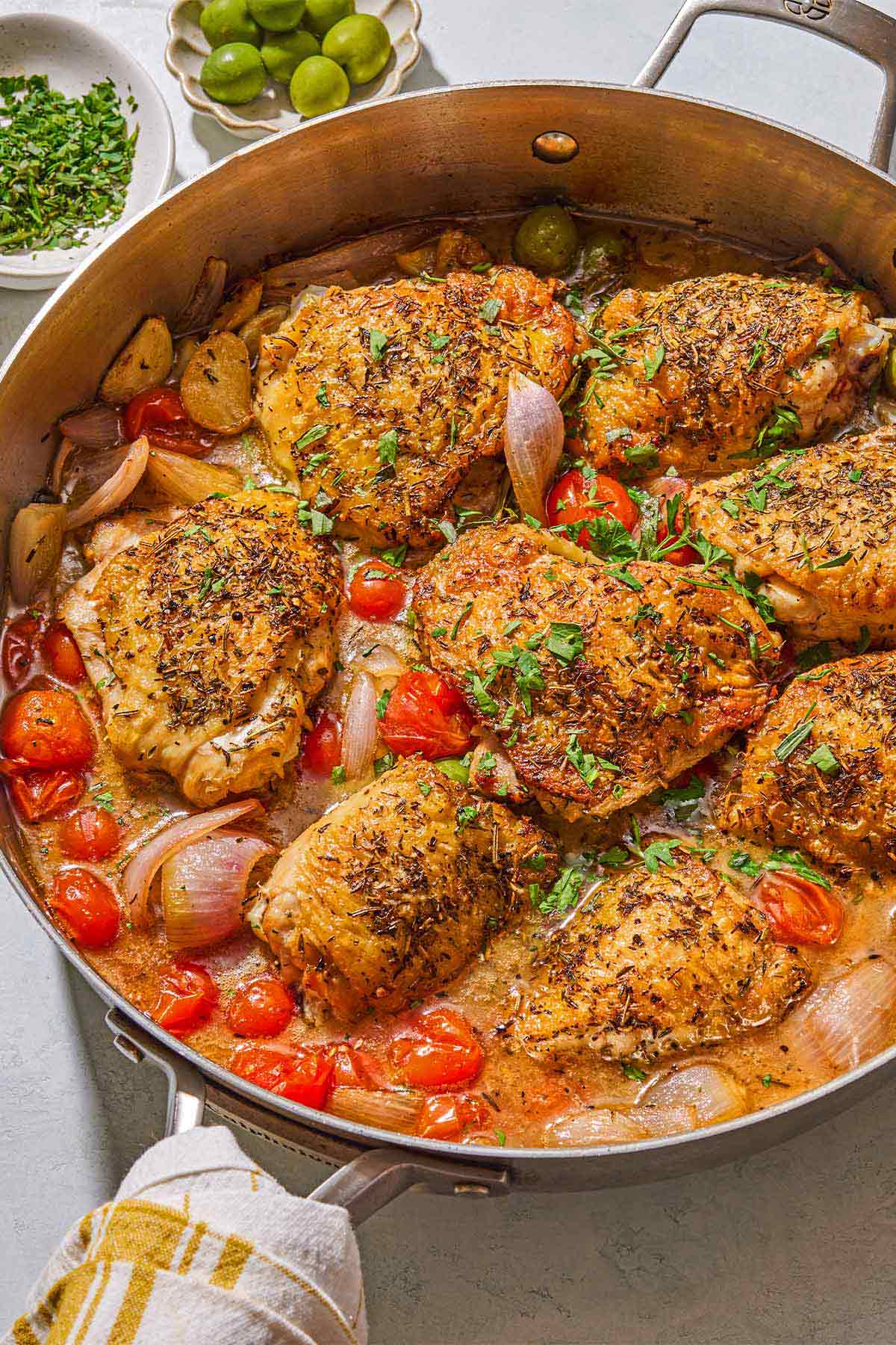 The chicken provencal in a skillet. Next to this are bowls of chopped herbs and green olives.