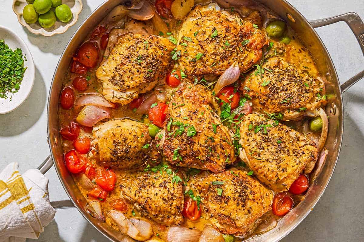 The chicken provencal in a skillet. Next to this are bowls of chopped herbs and green olives.