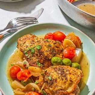 A serving of the chicken provencal on a plate. Next to this is a knife, a fork, a plate with French baguette pieces and a skillet with the rest of the recipe and a serving spoon.