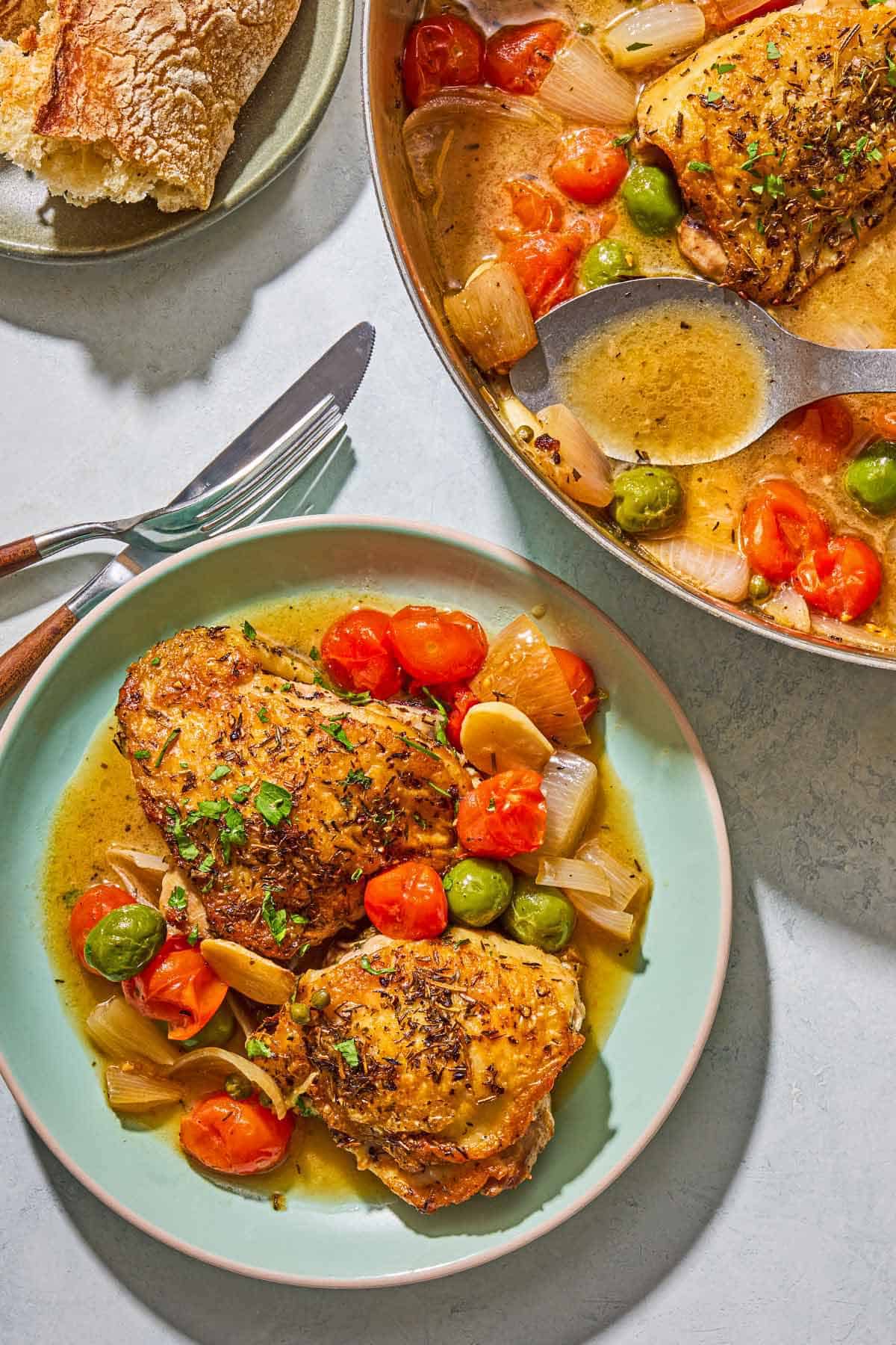 A serving of the chicken provencal on a plate. Next to this is a knife, a fork, a plate with French baguette pieces and a skillet with the rest of the recipe and a serving spoon.