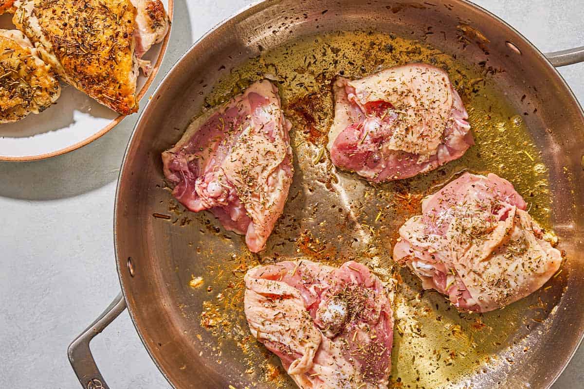 Four chicken thighs being browned in a skillet. Next to this is a plate with more browned chicken thighs.