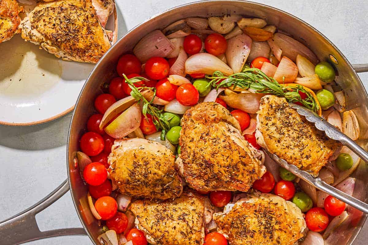 Chicken thighs for the chicken provencal being added to the pan with the herbs and vegetables with a pair of tongs. Next to this is a plate with the other more chicken thighs.