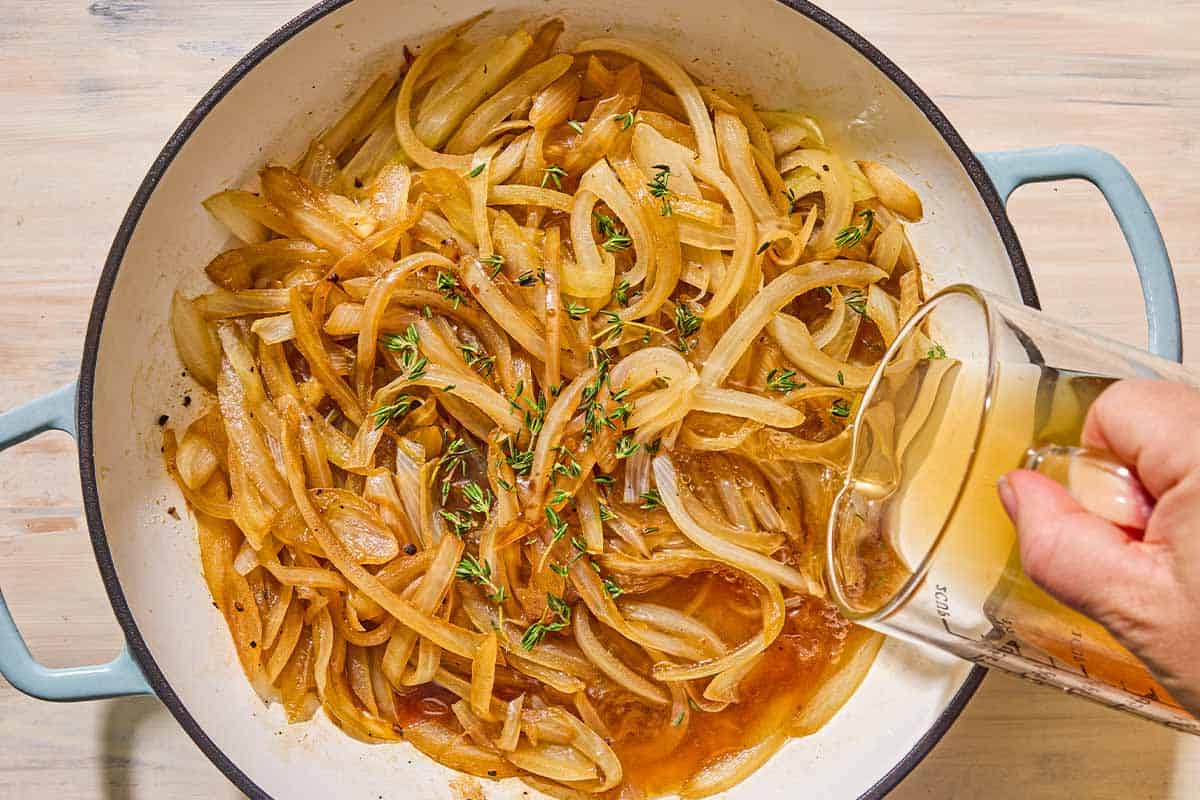 The beef broth being poured over the soften onions and thyme in a skillet.