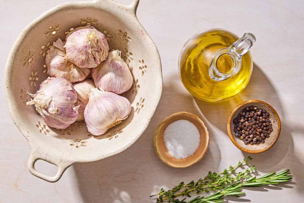 Ingredients for garlic confit including several bulbs of garlic, a bottle of olive oil, salt, pepper, fresh thyme and fresh rosemary.