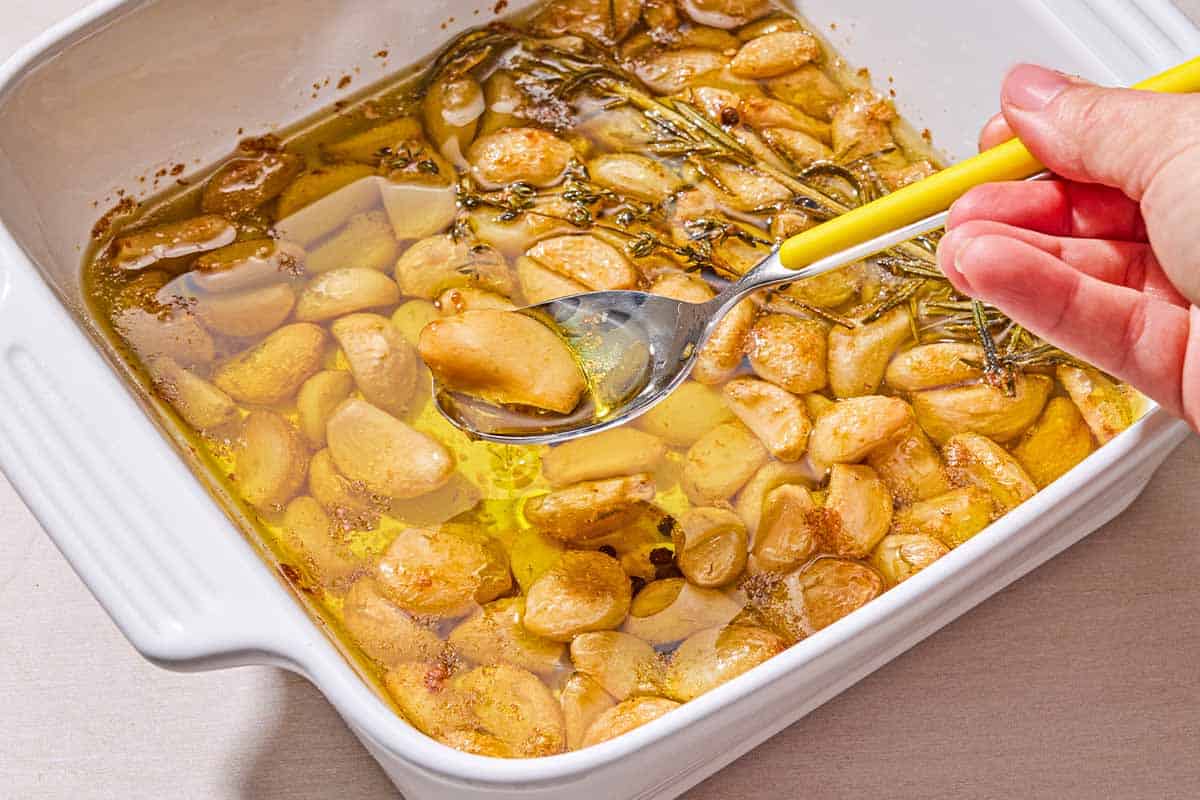 A clove of the garlic confit on a spoon being held over the rest of the confit in a baking dish.