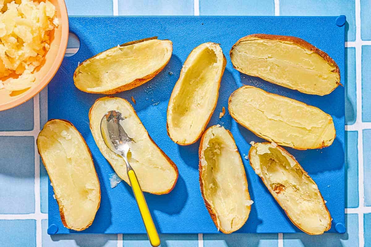 Several baked potato halves on a cutting board. Half have been scooped out. Next to the cutting board is a bowl with the scooped potato flesh.