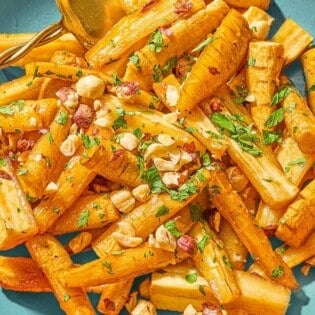 Roasted parsnips coated in a honey glaze and topped with hazelnuts and parsley in a serving bowl with a spoon.