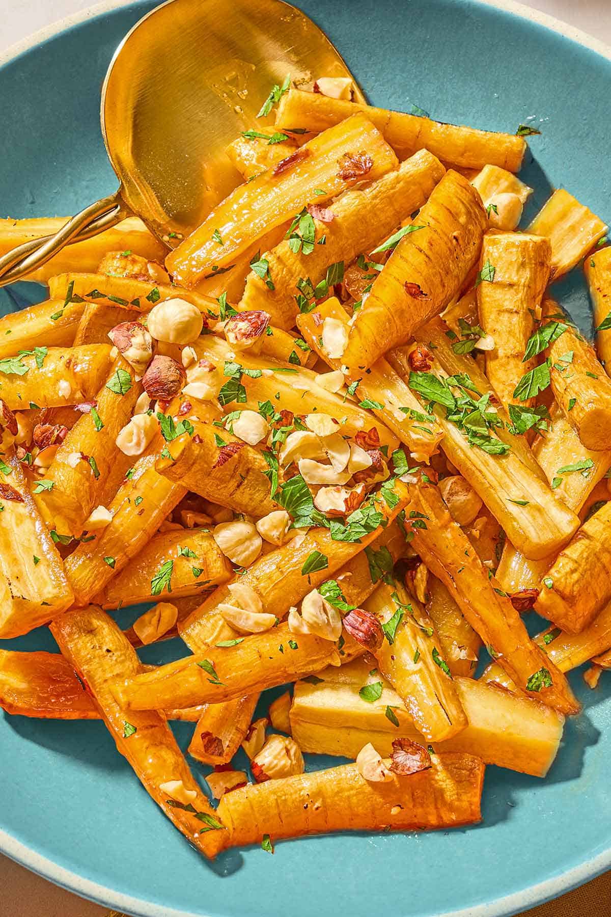 Roasted parsnips coated in a honey glaze and topped with hazelnuts and parsley in a serving bowl with a spoon.