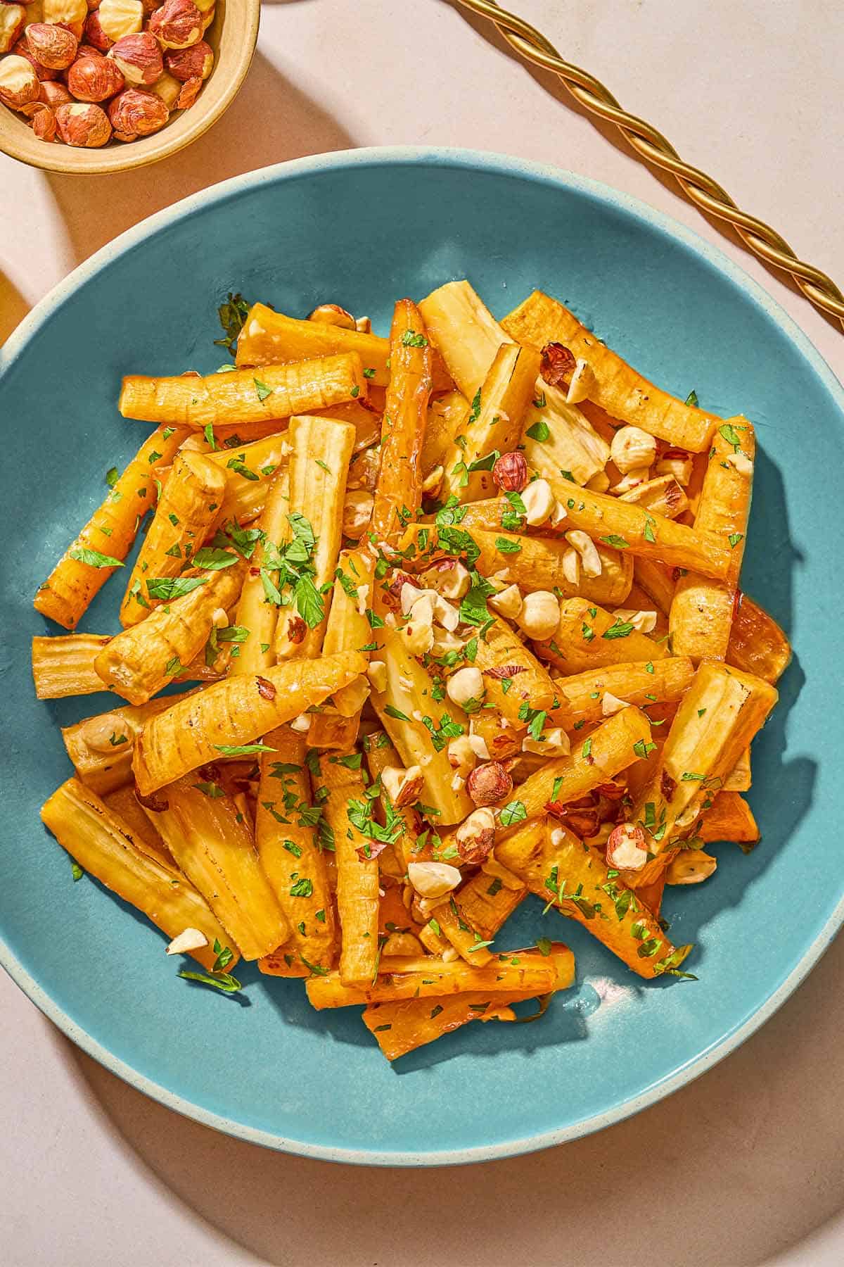 Roasted parsnips coated in a honey glaze and topped with hazelnuts and parsley in a serving bowl next to this is a bowl of roasted hazelnuts.