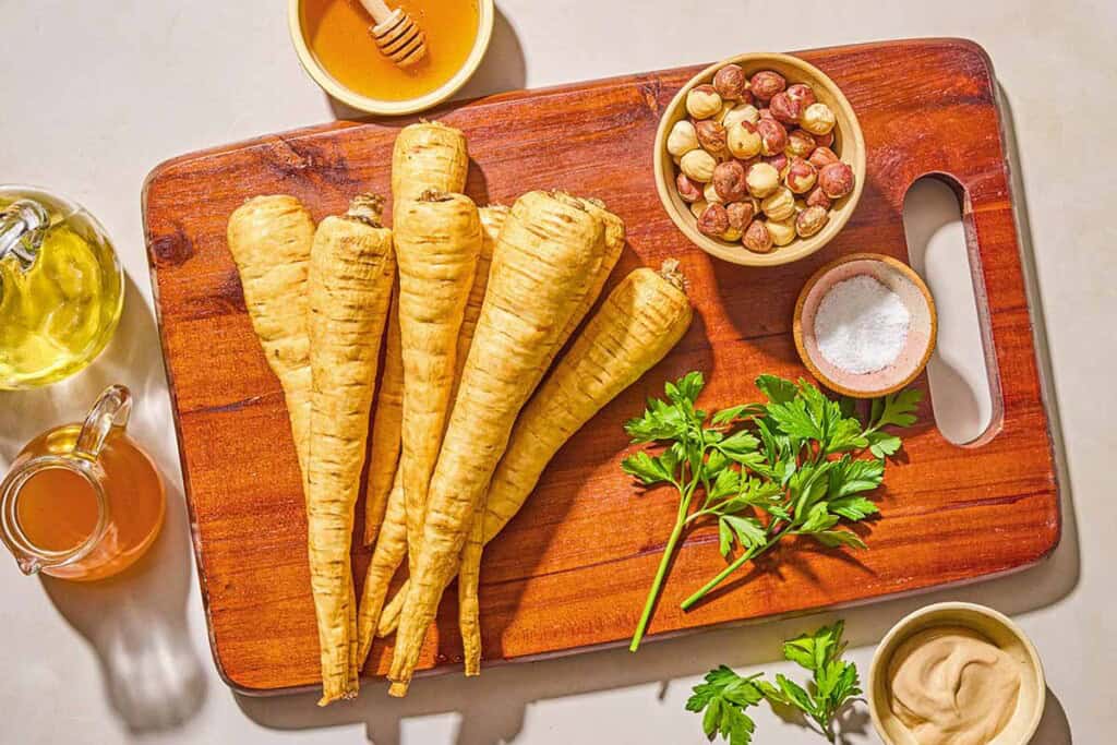 Ingredients for roasted parsnips including parsnips, olive oil, salt, honey, dijon mustard, apple cider vinegar, roasted hazelnuts, and italian parsley.