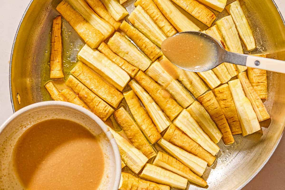 The honey mustard glaze being spooned over the roasted parnsips arranged in a single layer in a skillet.