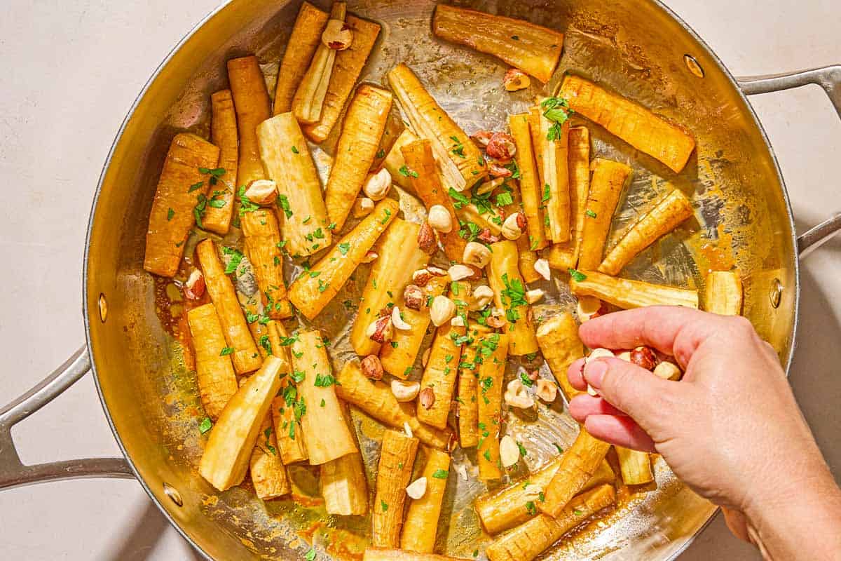 Roasted parsnips coated in a honey glaze and topped being topped with hazelnuts and parsley in a skillet.