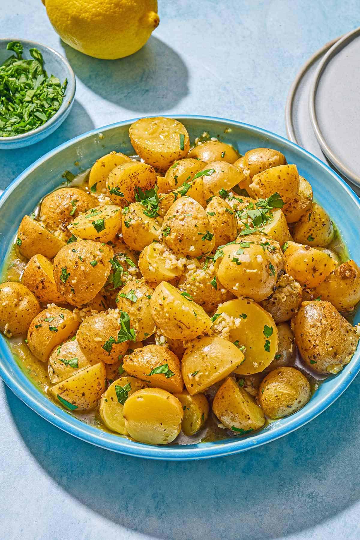 Roasted baby potatoes topped with chopped parsley in a serving bowl. Next to this is a bowl of chopped parsley, a lemon, and a stack of 2 plates.