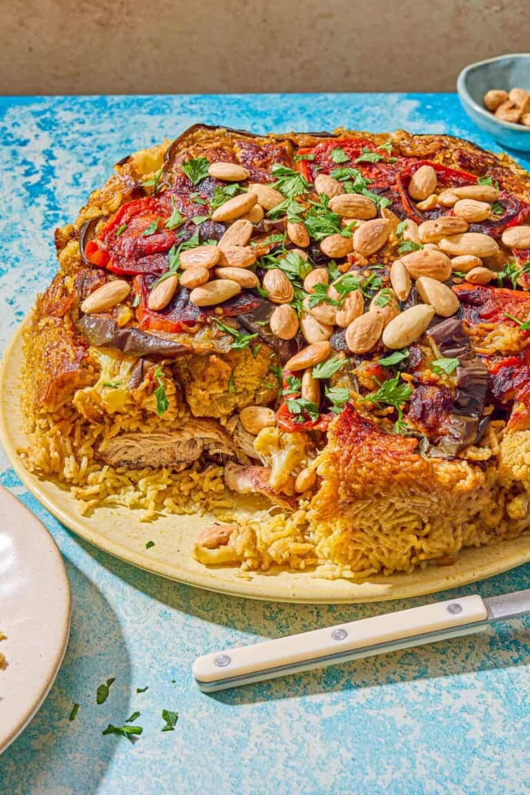 The maqluba on a platter next to a plate, a utensil and a small bowl of blanched almonds.