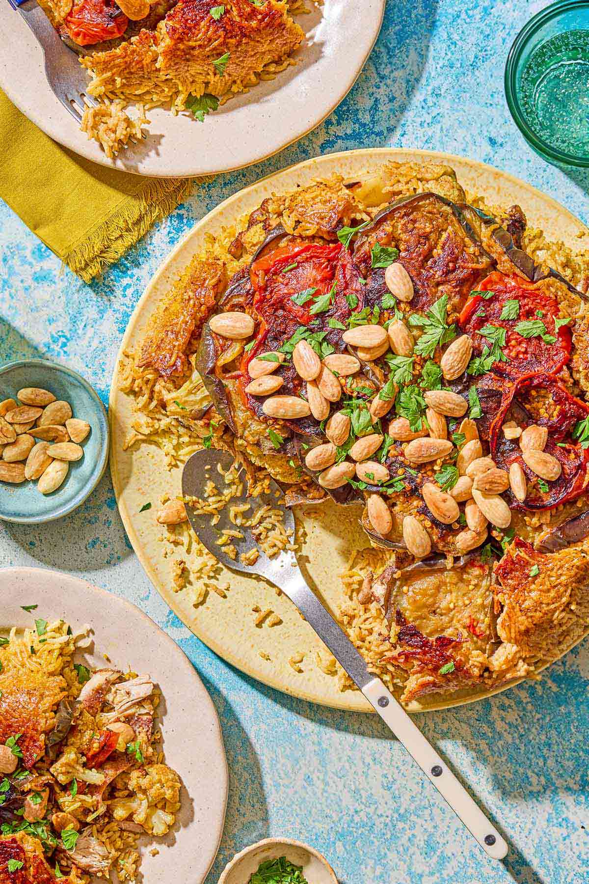 The maqluba on a platter with a serving spoon. Next to this is two servings of the recipe on 2 plates, small bowls of blanched almonds and chopped parsley and a glass of sparkling water.