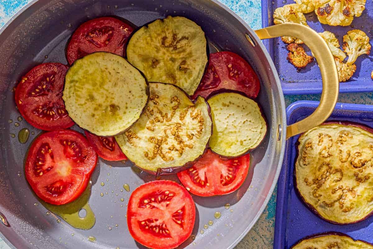A pot with roasted eggplant and tomatoes slices next to sheet pans with roasted cauliflower and roasted eggplant slices.