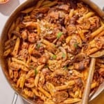 Mushroom pasta in a skillet with a wooden spoon. Next to this is a cloth napkin and bowls of chopped walnuts, red pepper flakes, parmesan cheese and parsley.