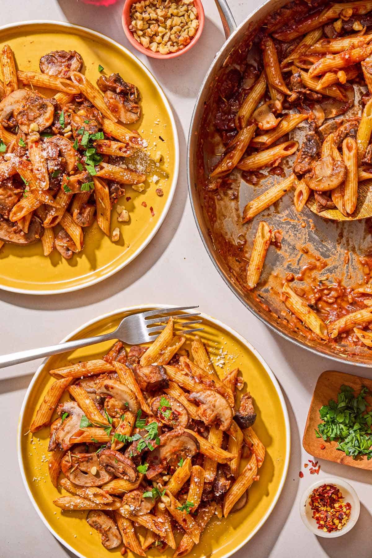 Two servings of mushroom pasta on plates, one with a fork. Next to these is the rest of the mushroom pasta in a skillet, and bowls of chopped walnuts, parsley and red pepper flakes.