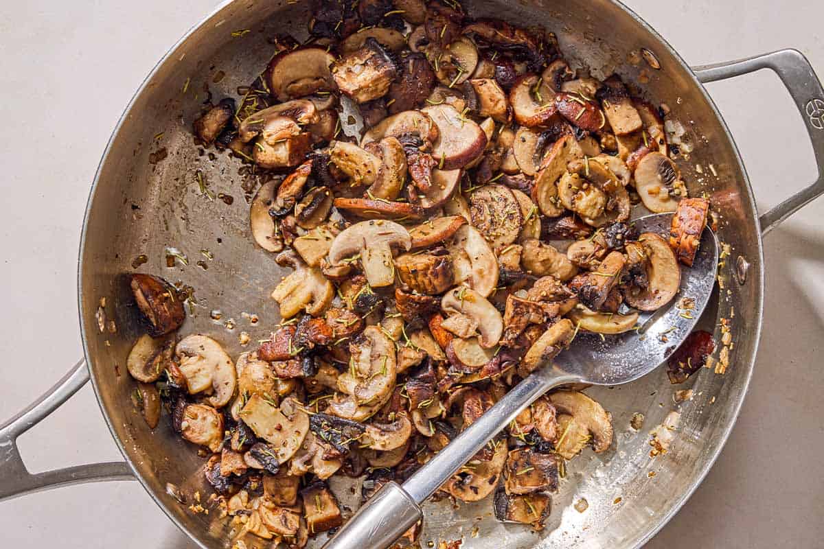 The sliced mushrooms, shallots, and garlic being sauteed in a skillet with a spoon.
