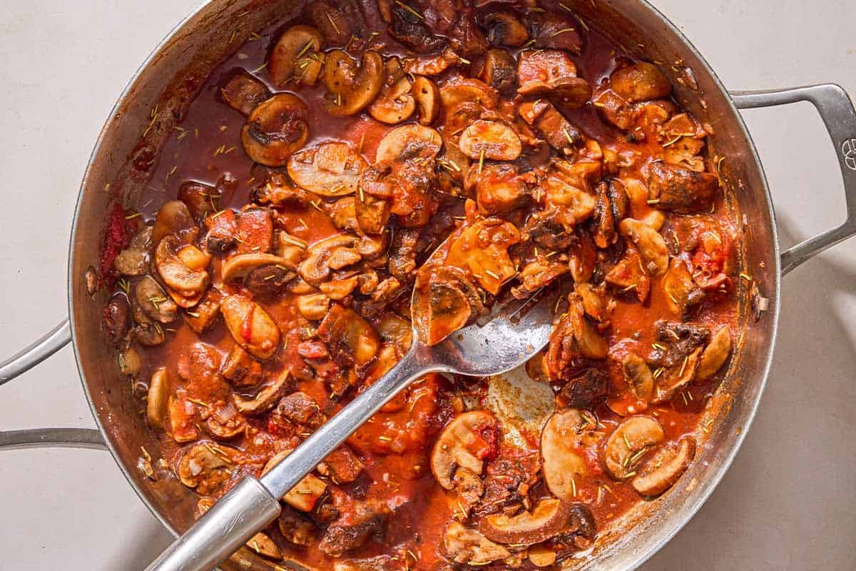 The mushrooms for the mushroom pasta simmering with the sauce in a skillet with a spoon.