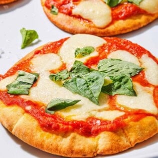 A close up of a pita pizza topped with fresh basil, with two more pizzas in the background.