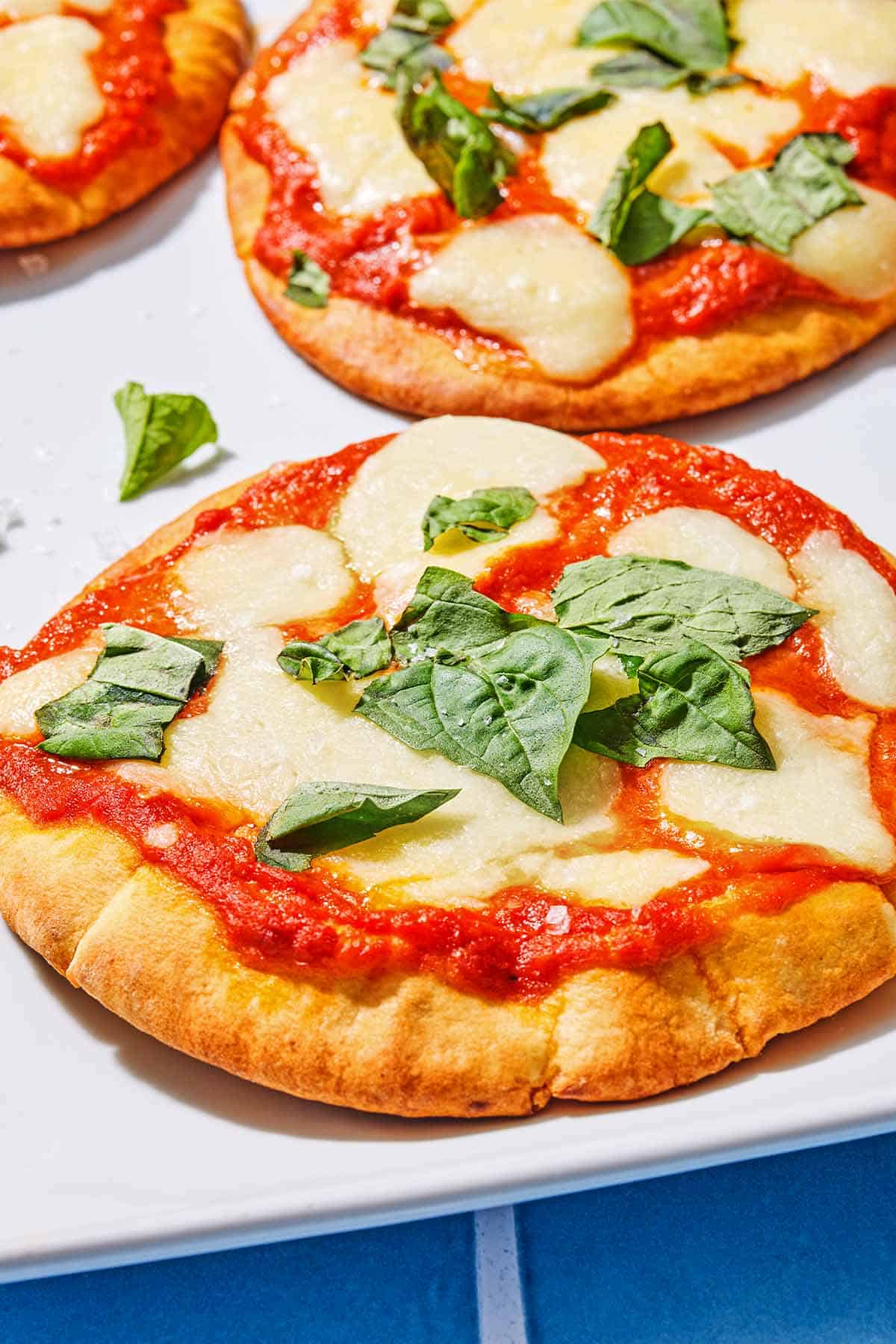 A close up of a pita pizza topped with fresh basil, with two more pizzas in the background.