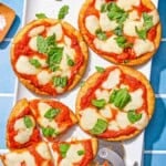 Four pita pizzas topped with fresh basil on a patter with a pizza slicer. One pita pizza is sliced into 4 pieces.