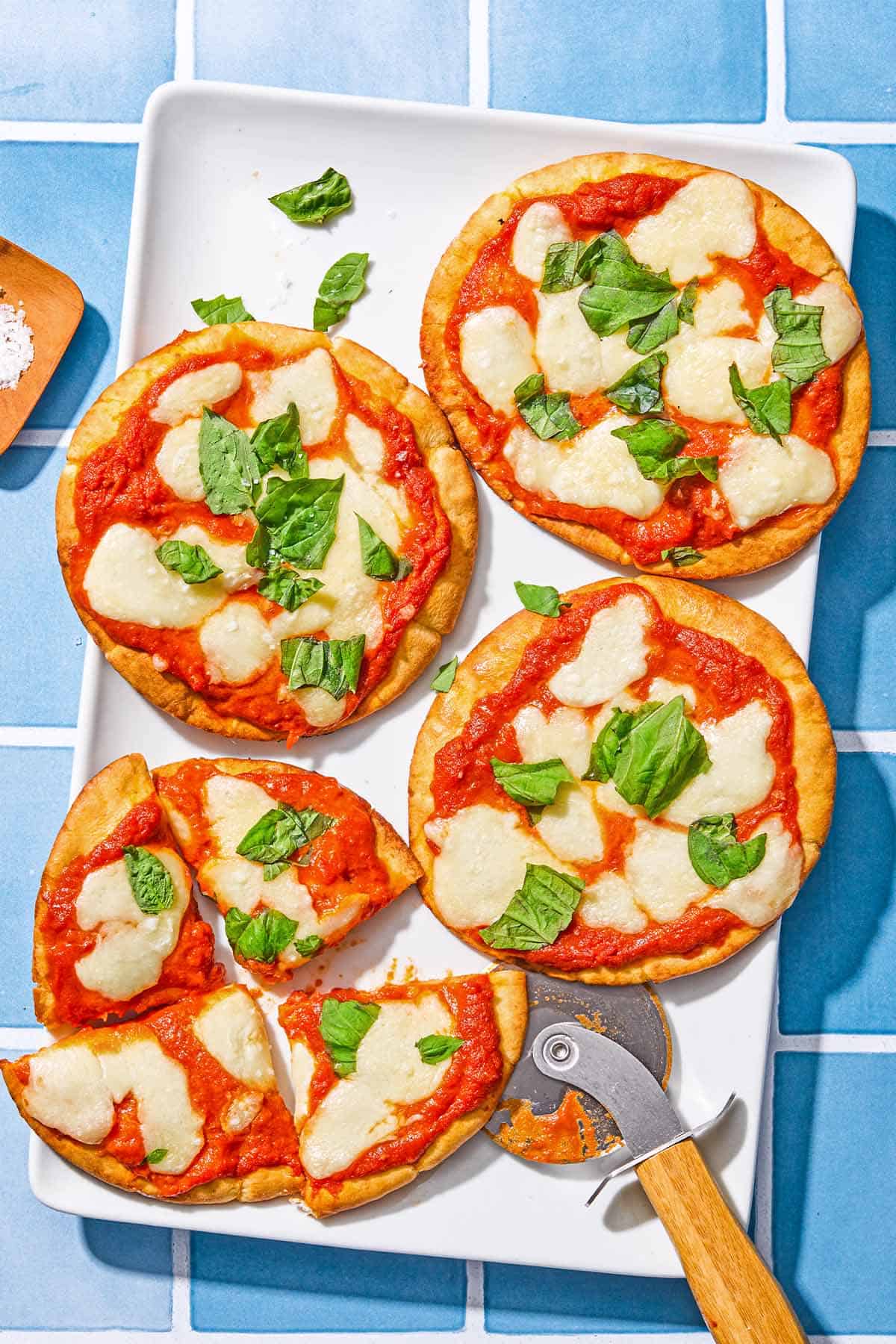 Four pita pizzas topped with fresh basil on a patter with a pizza slicer. One pita pizza is sliced into 4 pieces.